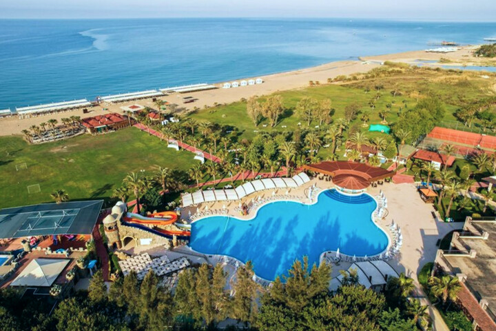 Luftaufnahme eines großen Schwimmbeckens mit Wasserrutsche, umgeben von Liegestühlen und Palmen an einem Strand mit Meer im Hintergrund.