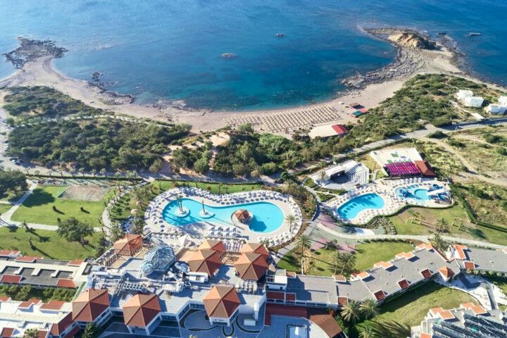 Luftaufnahme eines Resorts mit mehreren Pools, Sonnenschirmen, Gebäuden mit roten Dächern und einer Bucht mit Sandstrand und Felsen.