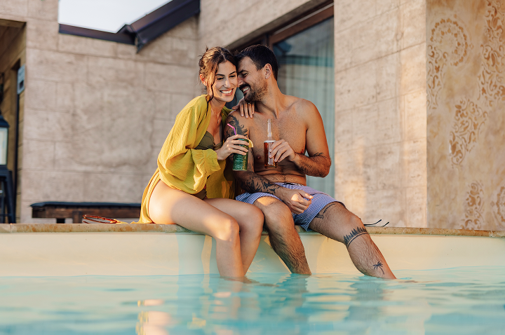 Paar sitzt am Poolrand, beide in Badebekleidung, halten Getränke und lehnen sich zärtlich aneinander.