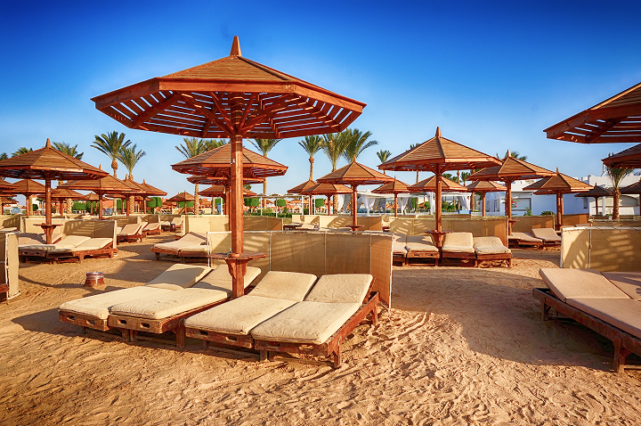 Mehrere hölzerne Sonnenschirme mit spitz zulaufenden Dächern über gepolsterten Liegen auf sandigem Boden, Palmen im Hintergrund.