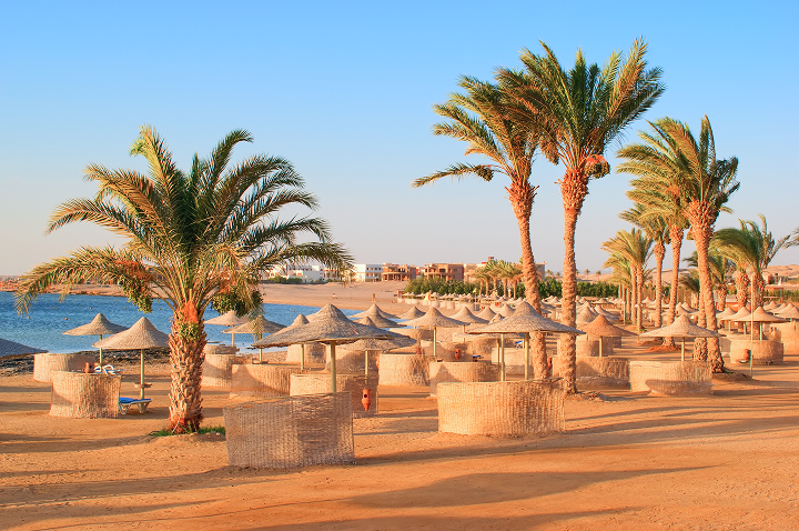 Strand mit Palmen und runden Strohhütten als Sonnenschutz, dahinter Meer und Gebäude am Horizont