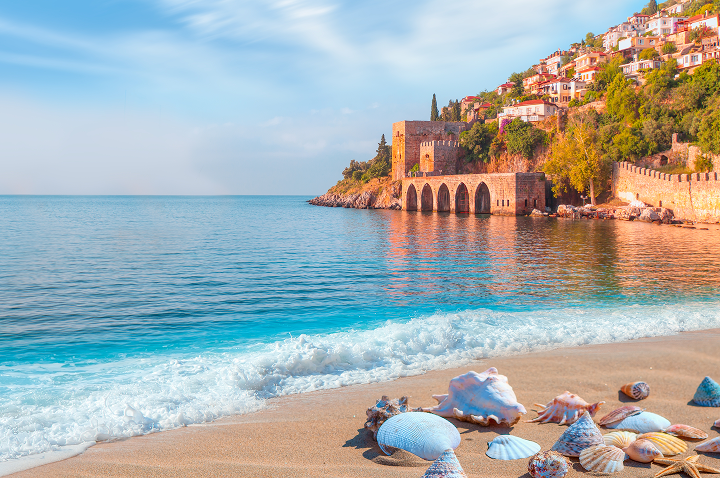 Strand mit Muscheln im Vordergrund, ruhiges Meer und bewaldeter Küstenabschnitt mit altem Steingebäude und Bögen.