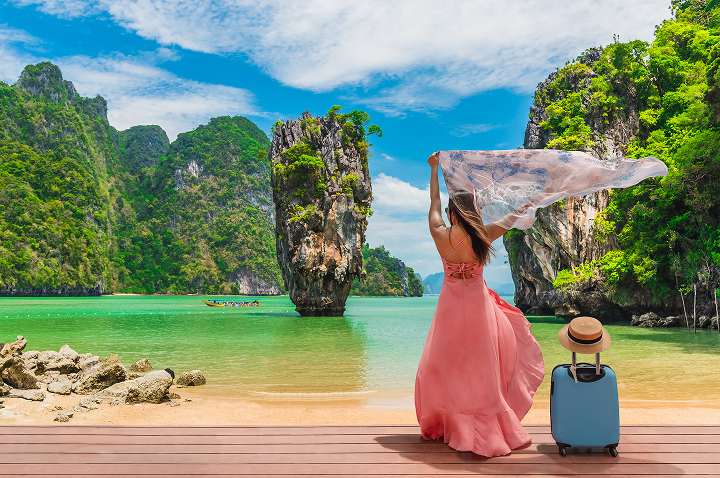 Auf einem Dock in Thailand steht eine Frau in einem pinken Kleid mit einem Koffer.