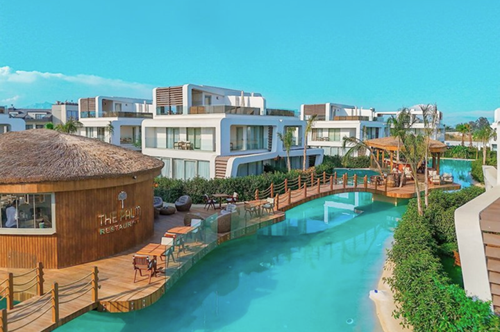 Moderner Hotelkomplex mit mehreren weißen Gebäuden, einem türkisfarbenen Wasserkanal und einem runden Restaurant mit Strohdach. Moderner Hotelkomplex mit mehreren weißen Gebäuden, einem türkisfarbenen Wasserkanal und einem runden Restaurant mit Strohdach.