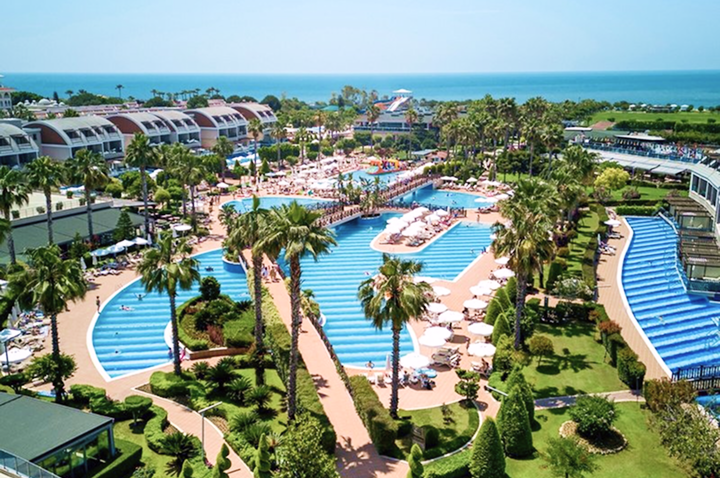Luftaufnahme einer weitläufigen Hotelanlage mit mehreren Pools, Palmen, Sonnenschirmen und Liegen, im Hintergrund das Meer.