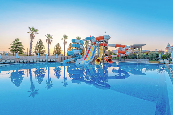 Wasserpark mit mehreren bunten Wasserrutschen und großem Schwimmbecken, umgeben von Liegestühlen und Palmen bei Sonnenuntergang.