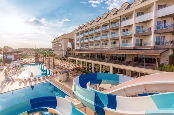 Hotelgebäude mit mehreren Stockwerken und Balkonen neben einem großen Pool mit Wasserrutschen und vielen Liegestühlen unter blauem Himmel.