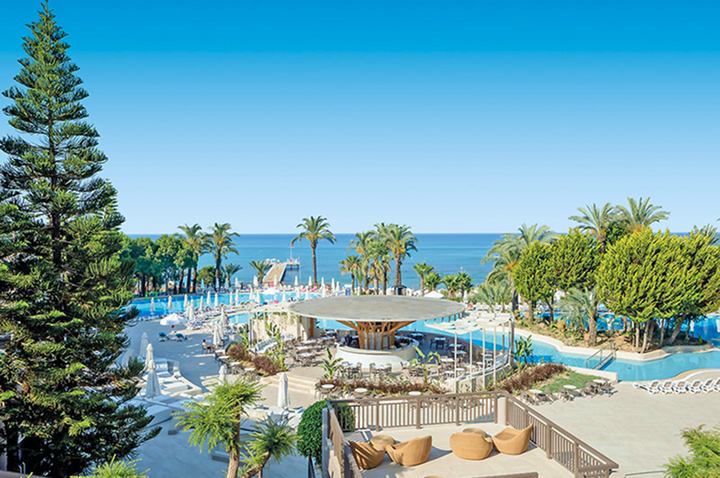 Blick auf eine weitläufige Poolanlage mit Palmen und Sitzbereichen vor dem Meer unter klarem Himmel.