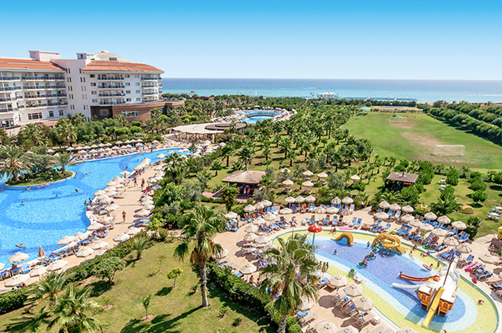 Blick auf ein Resort mit mehreren Pools, Liegen, Sonnenschirmen und Palmen, im Hintergrund das Meer.