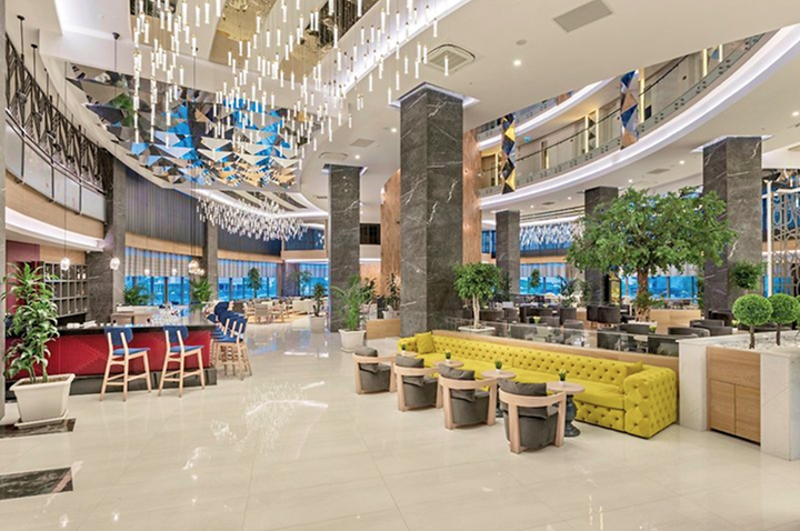 Großer, heller Hotellobbybereich mit gelbem Sofa, mehreren Sitzgruppen, grünen Pflanzen und moderner Deckenbeleuchtung.