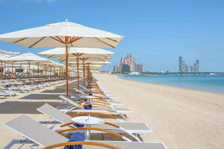 Strand mit Reihen von Liegestühlen und Sonnenschirmen, im Hintergrund das Atlantis-Hotel in Dubai am Meer.