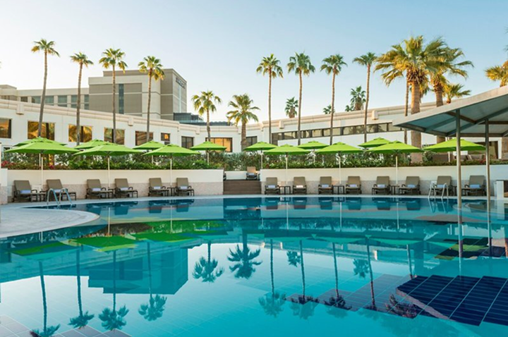 Hotelgebäude mit mehreren Stockwerken hinter einem Pool mit Liegestühlen und grünen Sonnenschirmen, umgeben von Palmen.