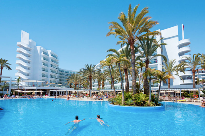 Großer Pool mit zwei Schwimmern, umgeben von Palmen und weißen Hotelgebäuden im Hintergrund.