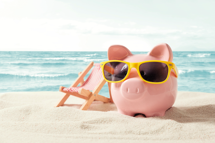 Sparschwein mit Sonnenbrille am Strand, neben einem Liegestuhl und einem Strandtuch.