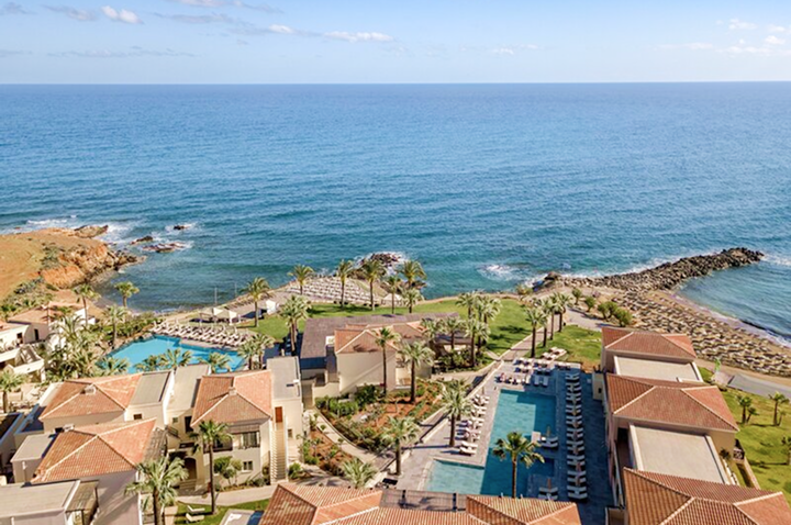 Luftaufnahme eines Resorts mit mehreren Gebäuden, Pools und Palmen an der Küste eines ruhigen Meeres.