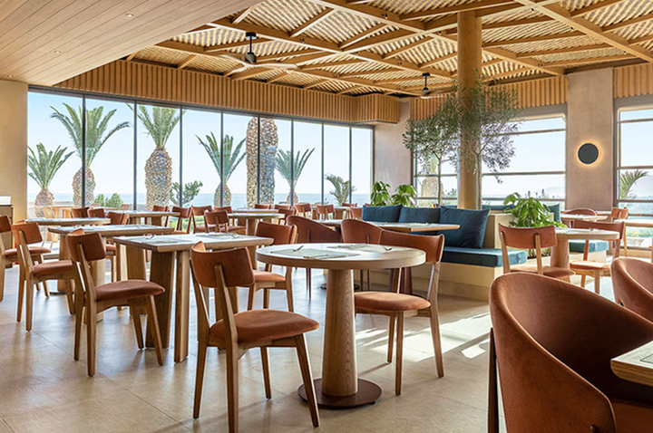 Helles Restaurant mit Holzbalkendecke, großen Fenstern, Palmenblick, Tischen mit braunen Stühlen und blauen Sitzbänken.
