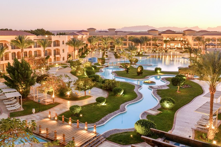 Hotelanlage mit geschwungenem Pool, beleuchteten Wegen, Palmen und mehrstöckigen Gebäuden bei Sonnenuntergang.