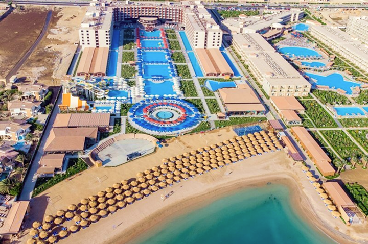 Luftaufnahme einer großen Hotelanlage mit mehreren Pools, Palmen und dicht gereihten Sonnenschirmen am Strand vor türkisfarbenem Wasser.