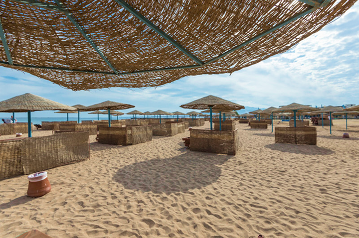 Strand mit mehreren Strohhut-Sonnenschirmen und windgeschützten Sitzbereichen aus geflochtenem Material auf Sand unter blauem Himmel mit Wolken Strand mit mehreren Strohhut-Sonnenschirmen und windgeschützten Sitzbereichen aus geflochtenem Material auf Sand unter blauem Himmel mit Wolken