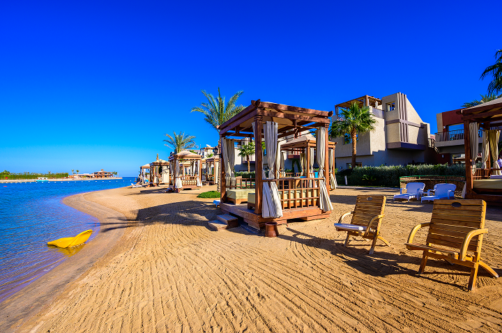 Strand mit mehreren Holzpavillons und Liegestühlen, Palmen und Gebäuden im Hintergrund unter klarem blauem Himmel.