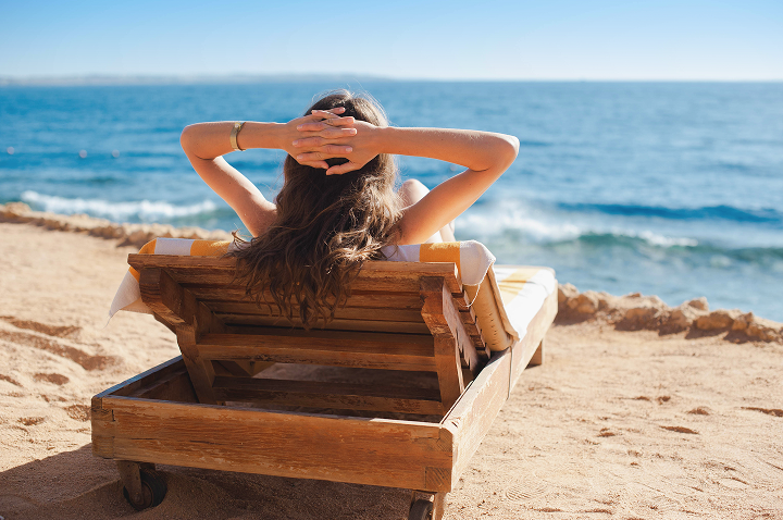 Eine Frau sitzt in einem Holzstuhl am Strand und genießt die frische Meeresbrise und die Sonne.