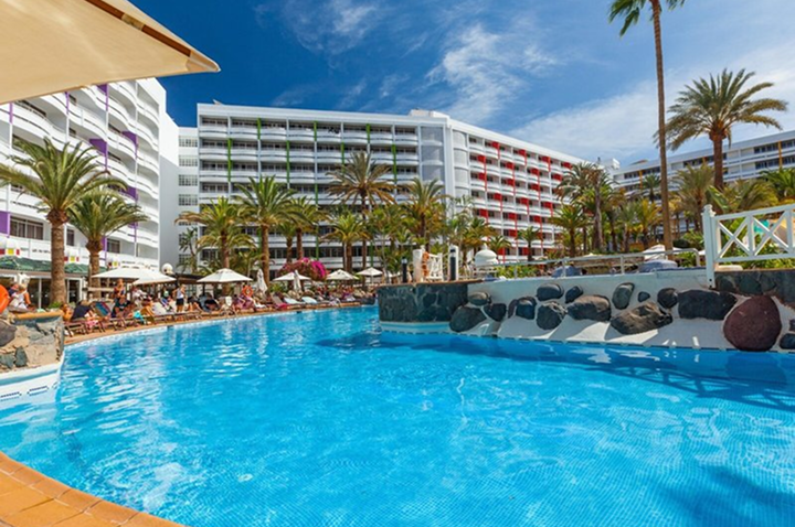 Großer Außenpool mit Sonnenschirmen und Liegestühlen vor einem mehrstöckigen Hotelgebäude und Palmen unter blauem Himmel.