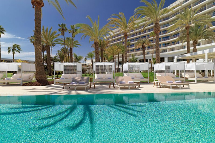 Hotelgebäude mit mehreren Stockwerken hinter einer Poollandschaft mit Liegen und Palmen. Hotelgebäude mit mehreren Stockwerken hinter einer Poollandschaft mit Liegen und Palmen.