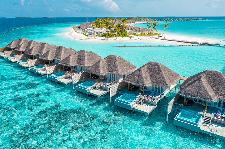 Luftaufnahme eines Resorts mit mehreren Reetdachhütten, umgeben von üppigem Grün und klarem Wasser.