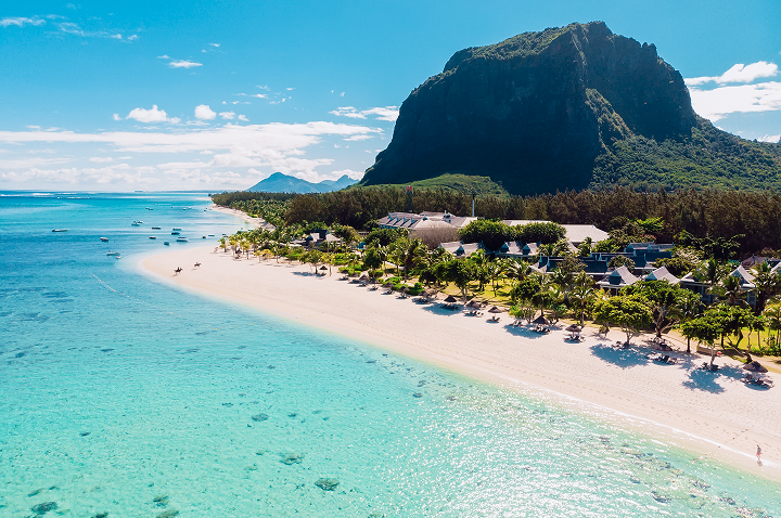 Strand mit türkisfarbenem Wasser, Palmen und Gebäuden am Fuß eines hohen, bewaldeten Bergs unter blauem Himmel mit Wolken.