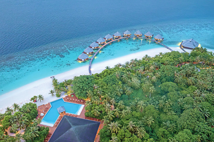 Luftaufnahme einer tropischen Insel mit Palmen, mehreren über Wasser gebauten Bungalows in Halbkreisform und einem Poolbereich am Strand.
