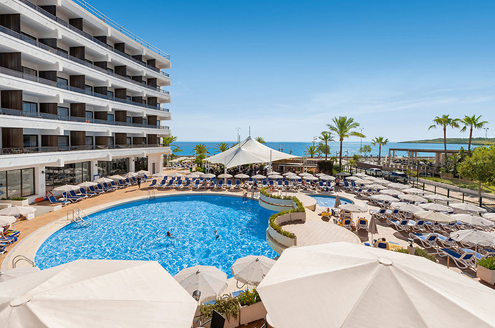 Hotel mit rundem Pool, Liegestühlen und Sonnenschirmen, Palmen und Meer im Hintergrund.