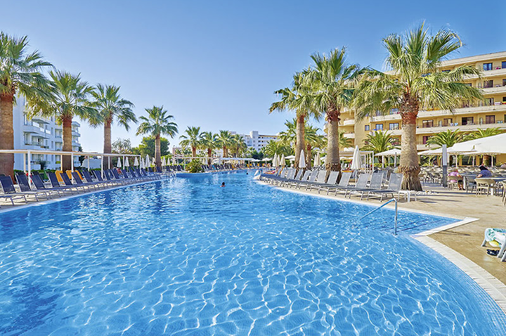 Großer Außenpool mit Palmen und Liegestühlen an beiden Seiten vor mehrstöckigen Hotelgebäuden