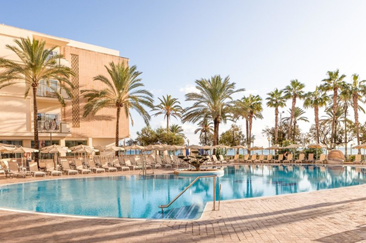 Großer Außenpool mit Liegestühlen und Sonnenschirmen vor einem mehrstöckigen Hotelgebäude und Palmen unter klarem Himmel.