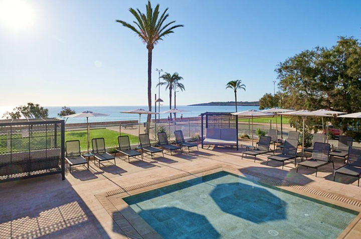 Poolbereich mit Liegestühlen und Sonnenschirmen vor Meerblick und Palmen unter klarem Himmel.