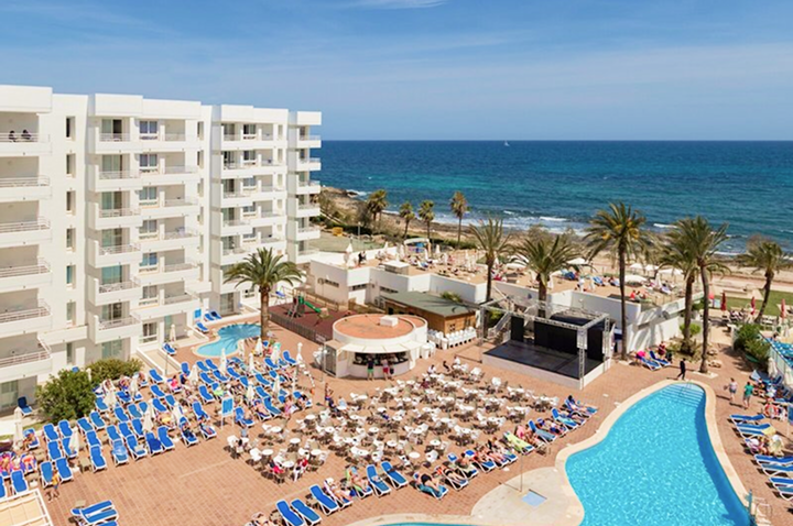 Hotelanlage mit mehreren Stockwerken, Pool, vielen Liegestühlen und Tischen auf der Terrasse sowie Strand und Meer im Hintergrund