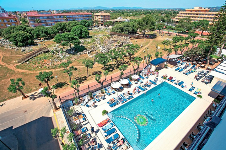 Hotelpool mit Liegestühlen und Sonnenschirmen neben einer archäologischen Ausgrabungsstätte und mehreren mehrstöckigen Gebäuden im Hintergrund.