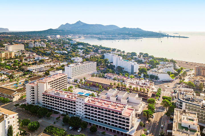 Luftaufnahme einer Küstenstadt mit mehreren mehrstöckigen Hotels, einem Strand und Bergen im Hintergrund.