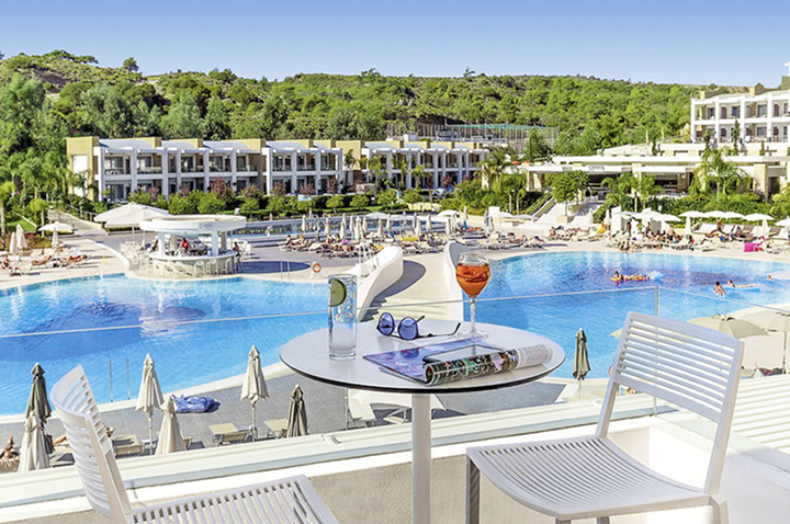 Blick von einer Terrasse mit Tisch und zwei Stühlen auf eine große Poollandschaft vor einem modernen Hotelkomplex mit mehreren Gebäuden und viel Grün.