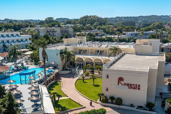 Großer Swimmingpool mit mehreren Palmen und strohgedeckten Pavillons vor einem weißen Hotelgebäude.