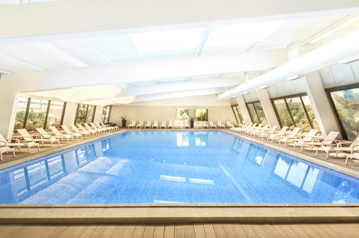 Innenraum eines Schwimmbads mit großem Becken, umgeben von Liegestühlen und großen Fenstern