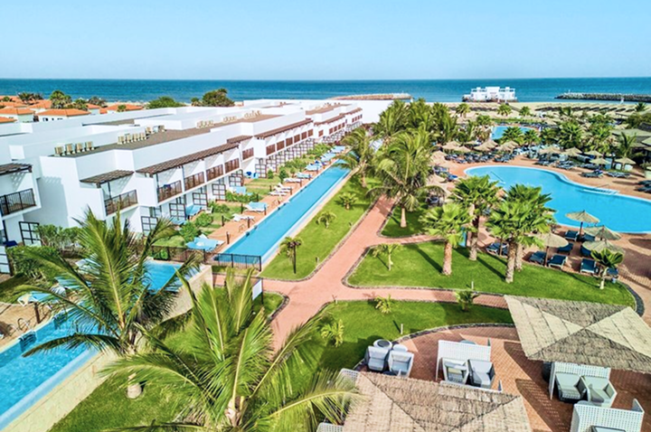 Blick auf ein Resort mit langem Pool, mehreren Gebäuden, Palmen und einem großen, geschwungenen Schwimmbecken am Meer.