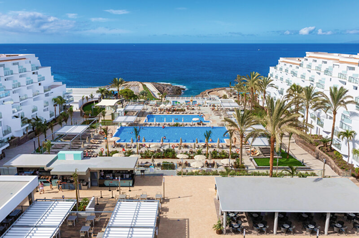 Blick auf ein Hotelresort mit mehreren Pools, Liegen und Palmen, im Hintergrund das offene Meer unter blauem Himmel.