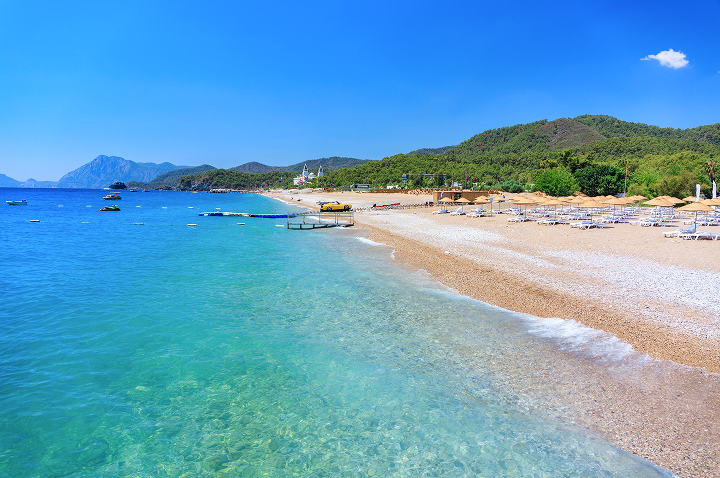 Klarer Strand mit Liegestühlen und Sonnenschirmen neben türkisfarbenem Meer und bewaldeten Hügeln unter blauem Himmel.