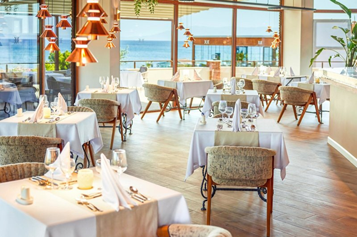 Restaurant mit mehreren eingedeckten Tischen, gepolsterten Stühlen und großen Fenstern mit Meerblick.