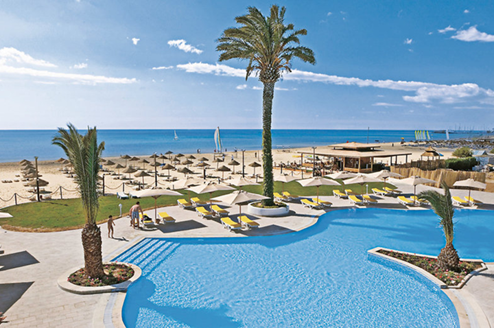 Großer Pool mit Palmen und Liegen, dahinter Sandstrand mit Sonnenschirmen und Meer unter blauem Himmel.